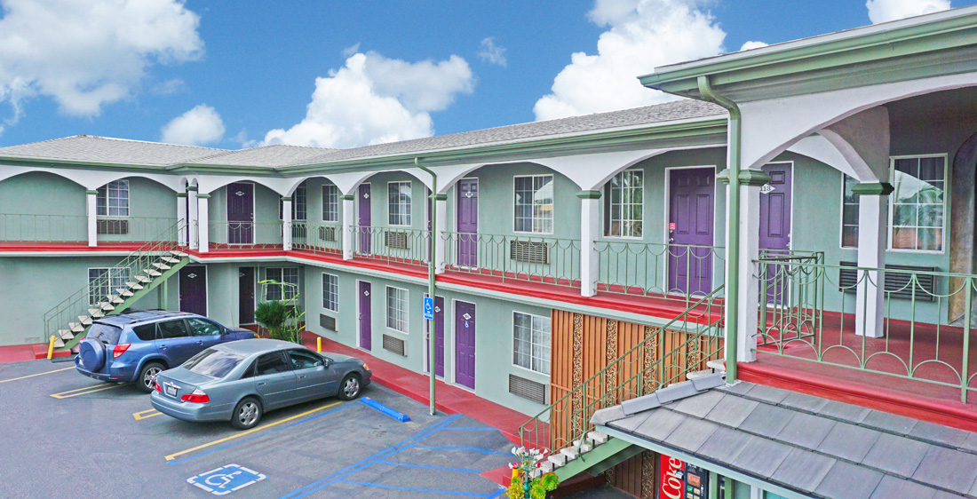 A two-story motel building with a green and red color scheme, a parking lot with two cars, and a blue sky with clouds.